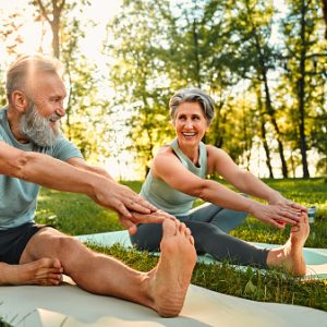 Flexible exercises for body. Sporty man and woman with grey hair stretching on yoga mats with hands to one leg during outdoors workout. Happy married couple with bare feet warming up together at park.